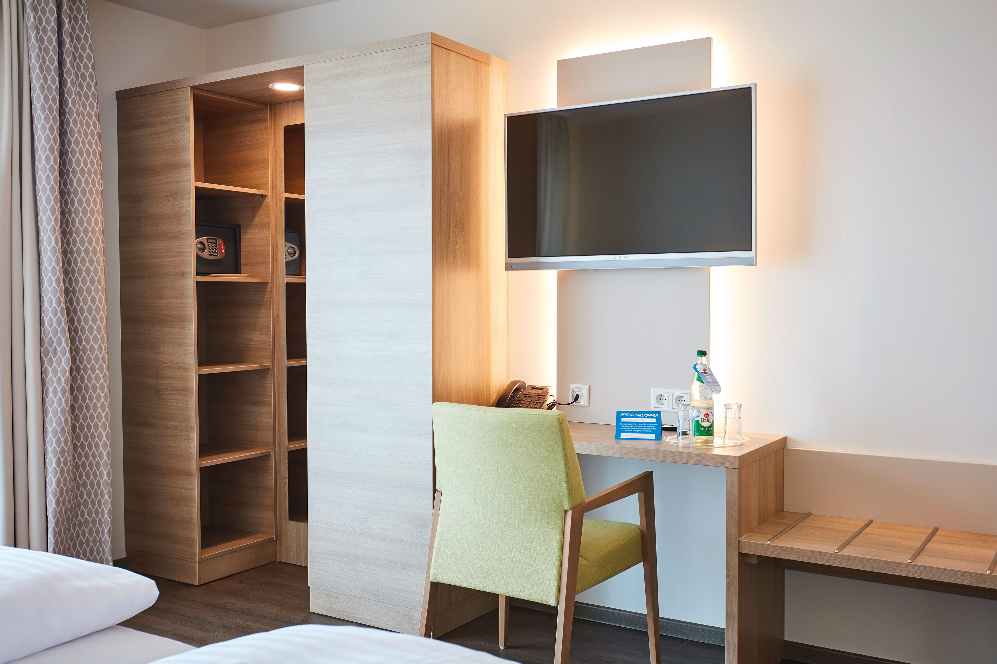 Das Bild zeigt den Schreibtisch im Doppelzimmer Komfort, über dem ein Fernseher angebracht ist, sowie links davon einen Kleiderschrank 