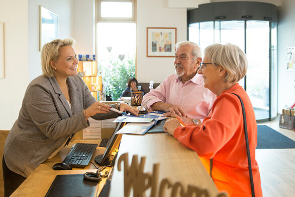 Das Bild zeigt die Geschäftsführerin des Vital Hotels, die an der Rezeption ein lachendes Paar herzlich empfängt