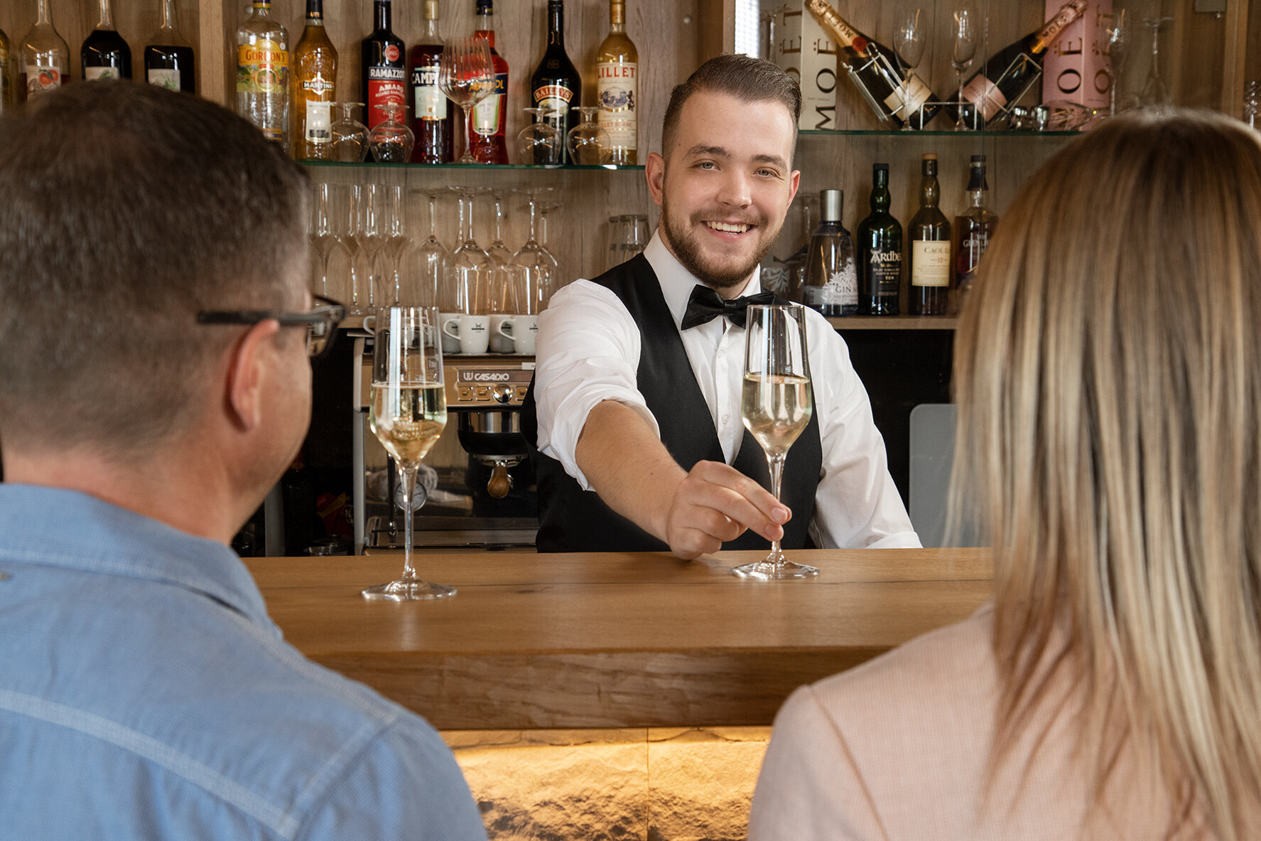 Das Bild zeigt den Barkeeper, der einem an der Bar sitzenden Paar zwei Gläser Sekt überreicht 