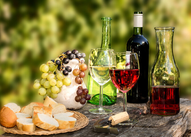 Das Bild zeigt Karaffen mit Wein, Weintrauben und ein Brett mit geschnittenem Baguette, die auf einem Stein im Freien dekoriert sind